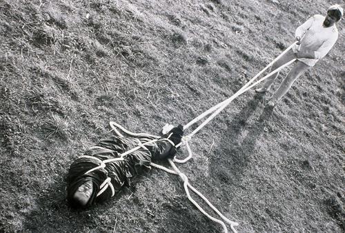 Foto einer Performance, eine Person liegt eingewickelt mit einem Seil auf dem Boden, eine andere Person hält das Seil fest 